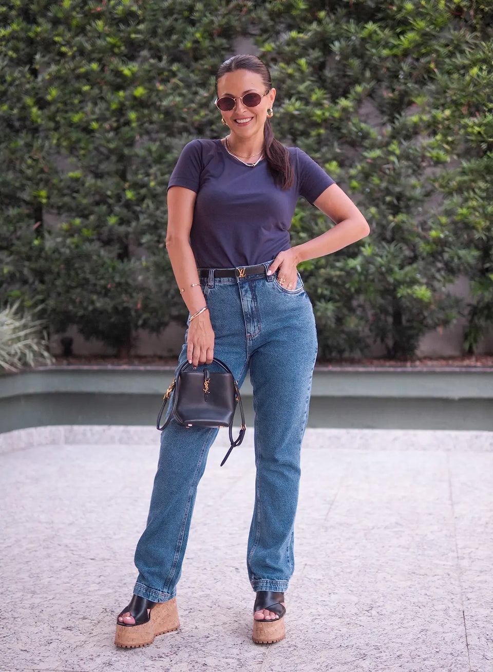 Mulher posando ao ar livre vestindo calça jeans azul e segurando bolsa de mão preta.