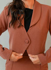 Mulher vestindo blazer marrom de botão único com blusa preta em fundo cinza.