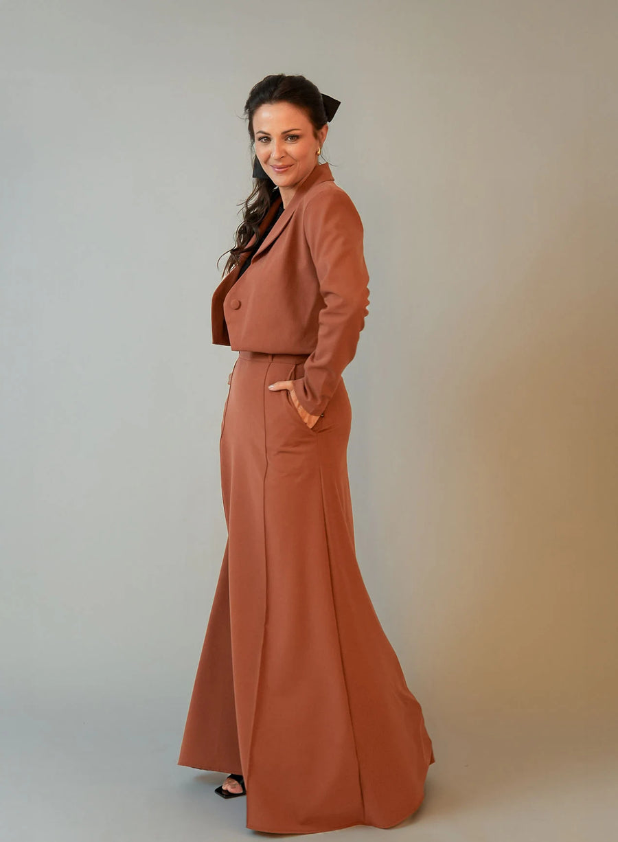 Mulher vestindo saia longa e blazer marrom em fundo neutro.