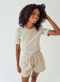 Mulher usa blusa tshirt bege e shorts, posando em ambiente minimalista.