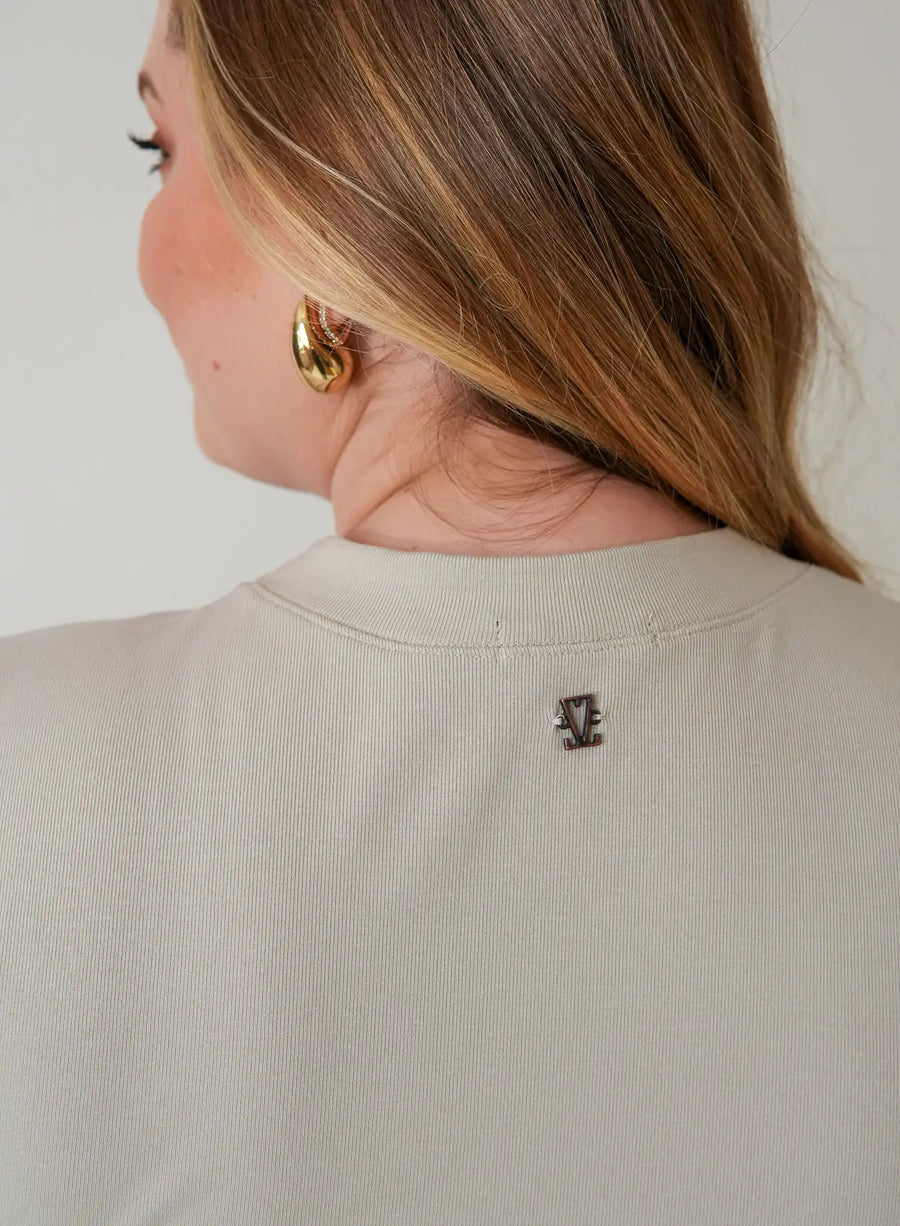 Parte de trás da blusa tshirt bege com pequeno pingente dourado representando logo da Elaz.