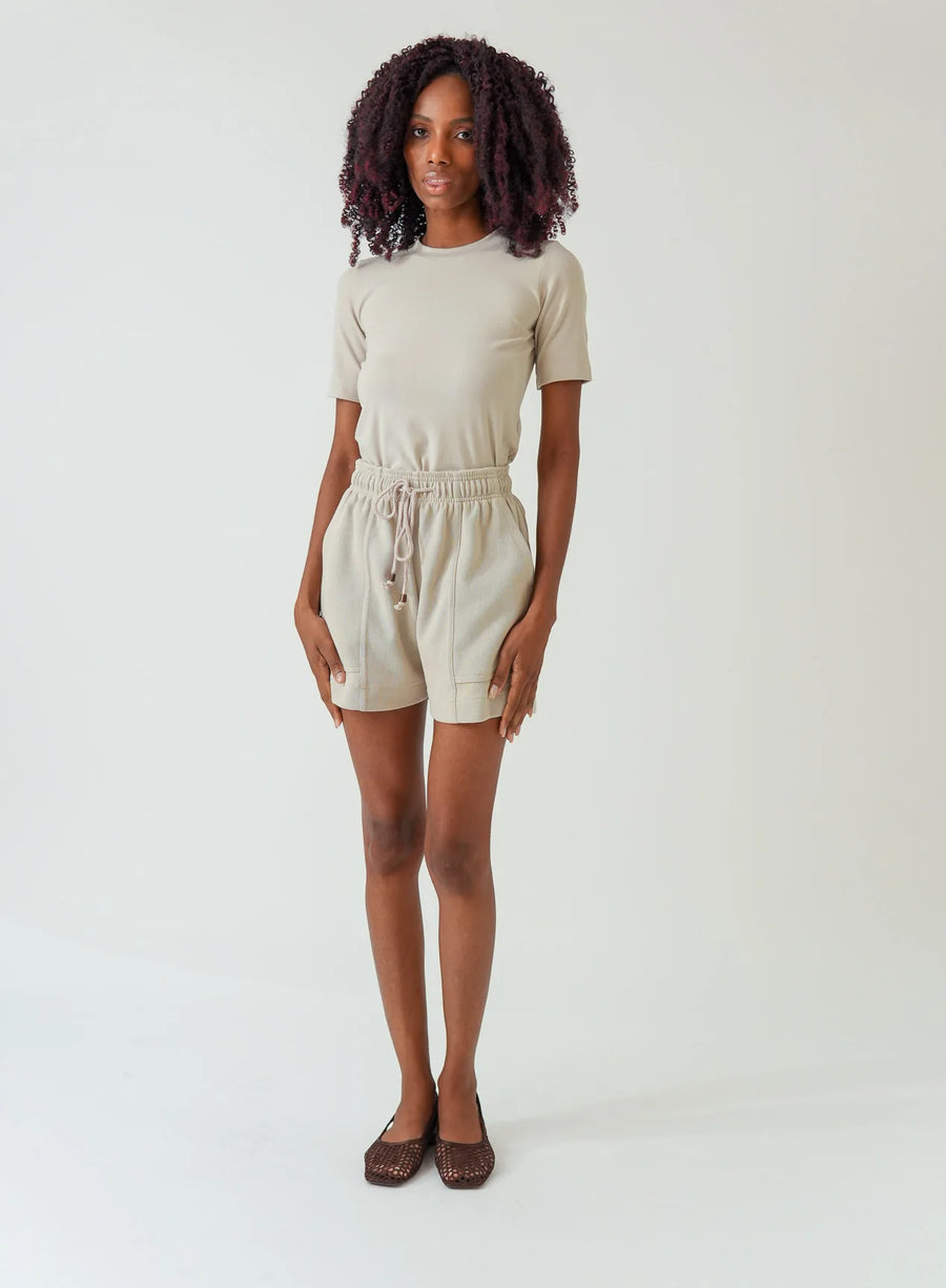 Modelo vestindo blusa tshirt bege e shorts da mesma cor em fundo neutro.