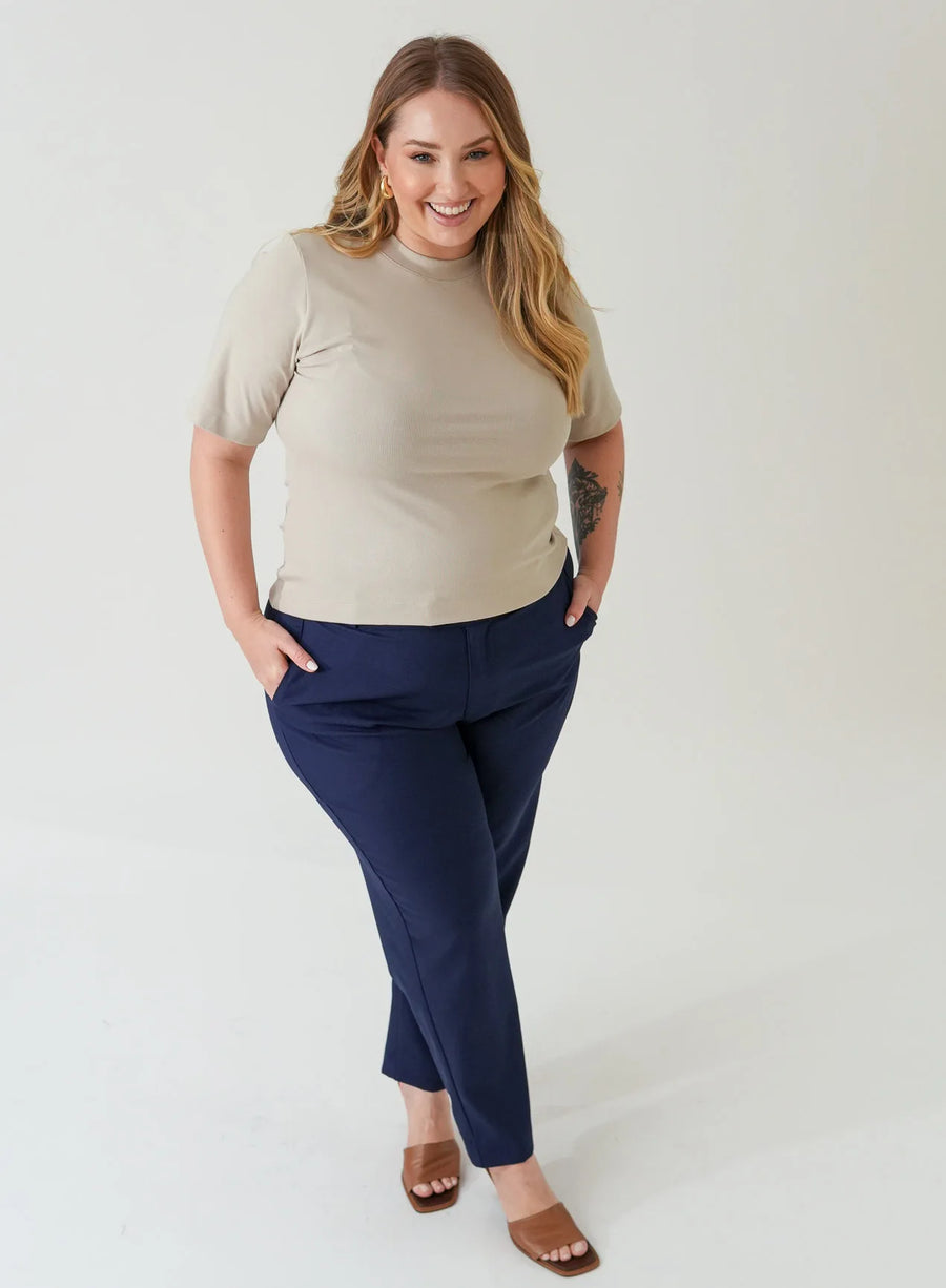 Mulher sorrindo com as mãos no bolso veste calça azul marinho. Ela também usa a blusa tshirt bege, ambas as peças são da Elaz.