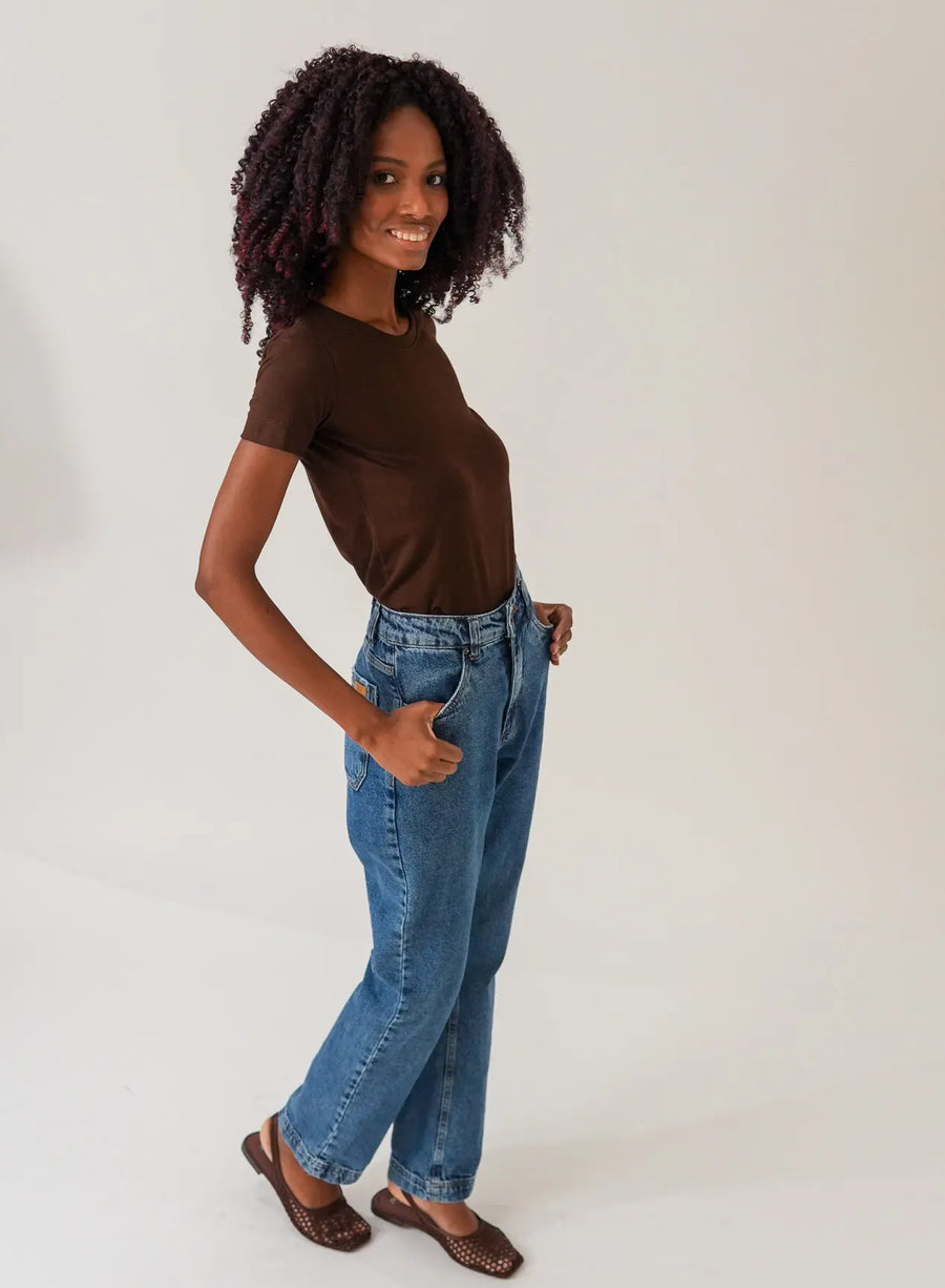 Mulher de lado posando com calça jeans azul e blusa thirt marrom em fundo neutro.