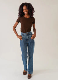 Mulher em fundo neutro vestindo blusa tshirt marrom e calça jeans azul.