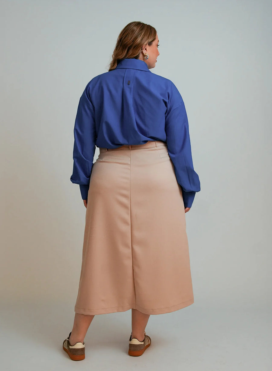 Modelo de costas em fundo neutro vestindo saia midi bege, par de tênis na cor marrom e camisa azul feminina.